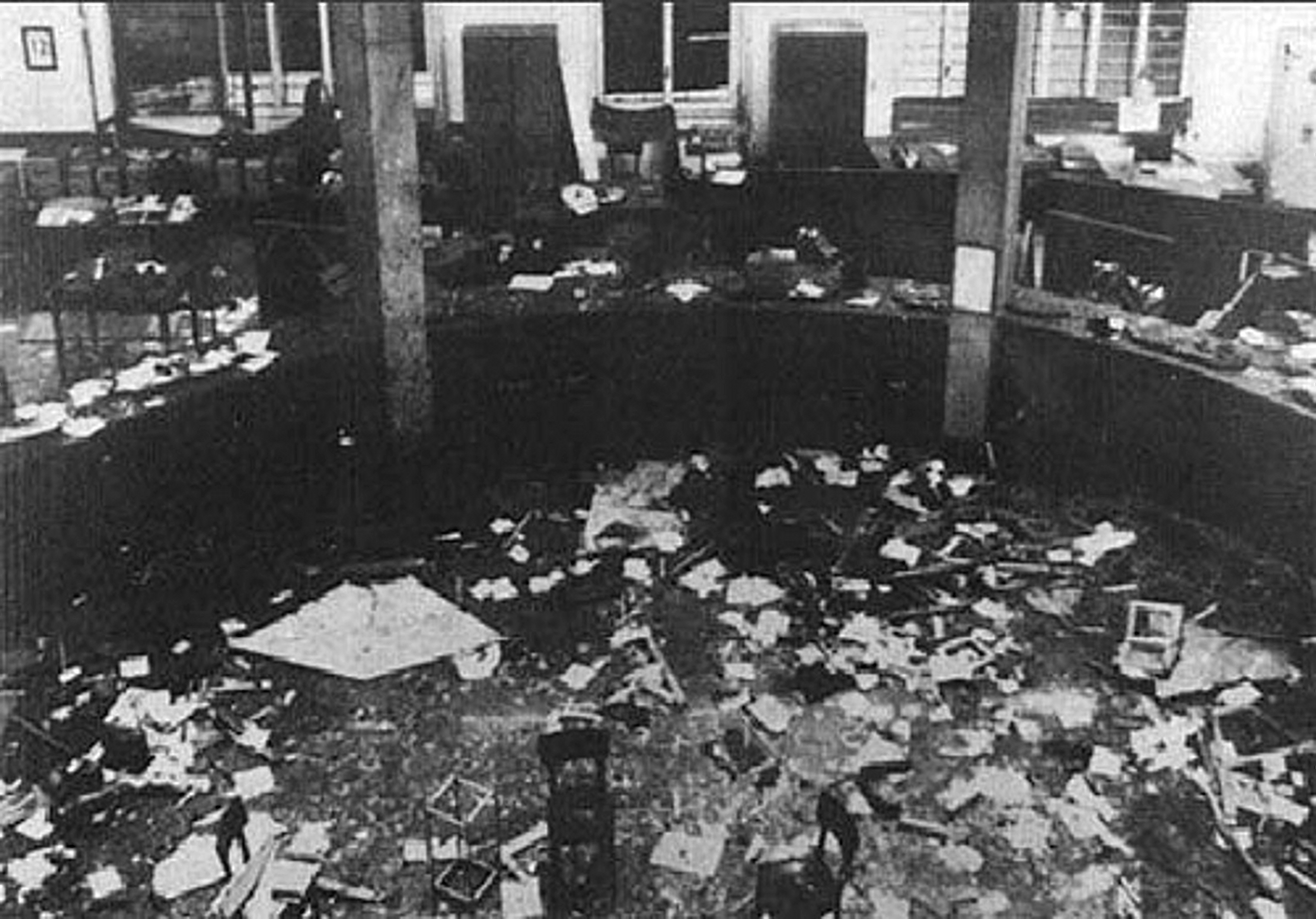 Immagine in bianco e nero dell'interno della Banca Nazionale dell'Agricoltura in seguito allo scoppio della bomba. Nell'immagine è visibile parte del pavimento e dei banchi disposti a semicerchio cosparsi di macerie, vetri rotti e carte.