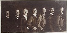 Augsta kontrasta fotosalona bilde, kurā septiņi vīrieši sastājušies līnijā viens aiz otra ieslīpi ar labo plecu pavērstu pret objektīvu, kreiso aiz priekšā stāvošā. Tie ir Apollo teātra direkcijas pārstāvji: Kristaps Koškins, Roberts Holcmanis, R. Millers, Ērihs Lauberts, Gustavs Žibalts, Reinholds Veics, Arveds Bergamanis.
