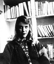 A black-and-white photo of a Caucasian woman with shoulder-length hair. She is seated facing the camera, wearing a sweater, with bookshelves behind her.