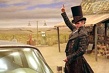 The character Count Olaf, played by Jim Carrey, sports an idyllic top hat and black-striped overcoat, seen from his thighs and up. Behind him shows a bleak cornfield on the left and a gas station on the right. The station has a red-and-white striped awning and looks worn. Olaf faces to the driver's side window of the car, and he positions his right hand as if he's pointing at something above him, while concentrating his eyes on the window and holding car keys in his left hand.