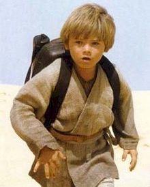 Un băiat blond cu haină gri și purtând la spate un rucsac merge prin deșert.