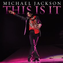 A picture of a man with dark hair pointing with his right hand to his right. The man is wearing a black jacket with a red shirt and black pants. Behind him, there is a black background with the words "MICHAEL JACKSON" in white print and, underneath it, "THIS IS IT" written in pink.