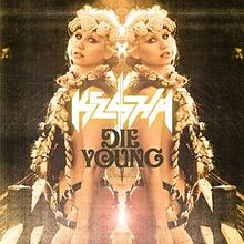 A Caucasian female with blond braids and a floral diadem. She is sparsely clothed in a suit of leather strips with much skin expose. Back facing a mirror, her reflection is symmetrically depicted with the words "Kesha" and "Die Young" appearing in the center.