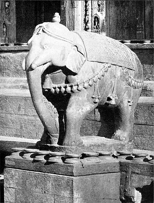 Black and white photograph of an statue of an elephant on a plinth. The background is a wall with some decoration near the top of the image.