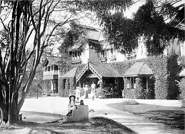 Black and white photograph of a large two storey house in the country. Four women stand in the background, by the front door. A girl sits on steps in the foreground, in the shade of a tree.
