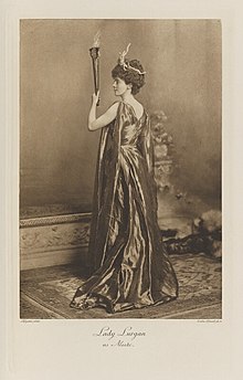 Black-and-white photograph of a standing woman richly dressed in an historical costume with her back to the viewer, holding a flaming torch and wearing a crown