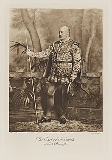 Black-and-white photograph of a standing man richly dressed in an historical costume with a staff, sword, hat and short cloak