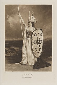 Black-and-white photograph of a standing woman richly dressed in an historical costume with a huge shield, very long spear, and a large helmet with feathers reaching up