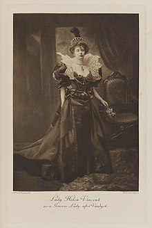 Black-and-white photograph of a standing woman richly dressed in an historical costume with a tiara and a black feather plume on top of her head