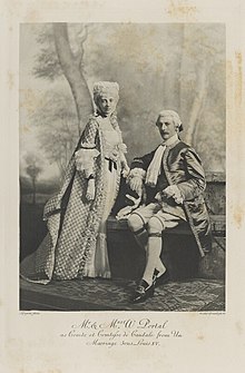 Black-and-white photograph of a standing woman and seated man richly dressed in historical costumes