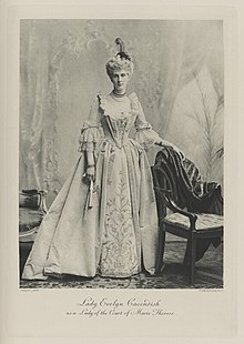 Black-and-white photograph of a standing woman richly dressed in an historical costume with a feather coming out of the top of her hat and a fan
