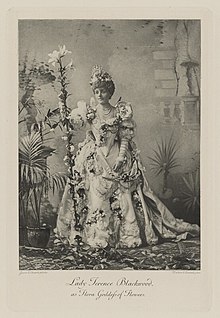 Black-and-white photograph of a standing woman richly dressed in an historical costume decorated all over with flowers