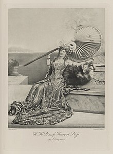 Black-and-white photograph of a woman seated on some steps and richly dressed in an historical costume, leaning back against the steps, holding an enormous fan, with feathers in her hair