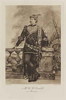 Black-and-white photograph of a standing man richly dressed in an historical costume in a tunic and tights