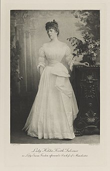 Black-and-white photograph of a standing woman richly dressed in an historical costume