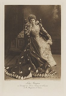 Black-and-white photograph of a standing woman richly dressed in an historical costume