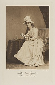 Black-and-white photograph of a seated woman richly dressed in an historical costume with her hair down and holding a book