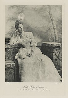 Black-and-white photograph of a seated woman richly dressed in an historical costume with a white feather plume in her hair and a fan