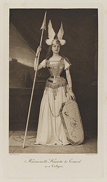 Black-and-white photograph of a standing young woman dressed in armor with a helmet, shield and spear